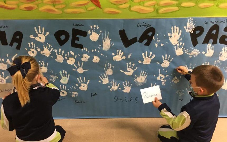 Two pupils write on a Peace Day mural covered in handprints