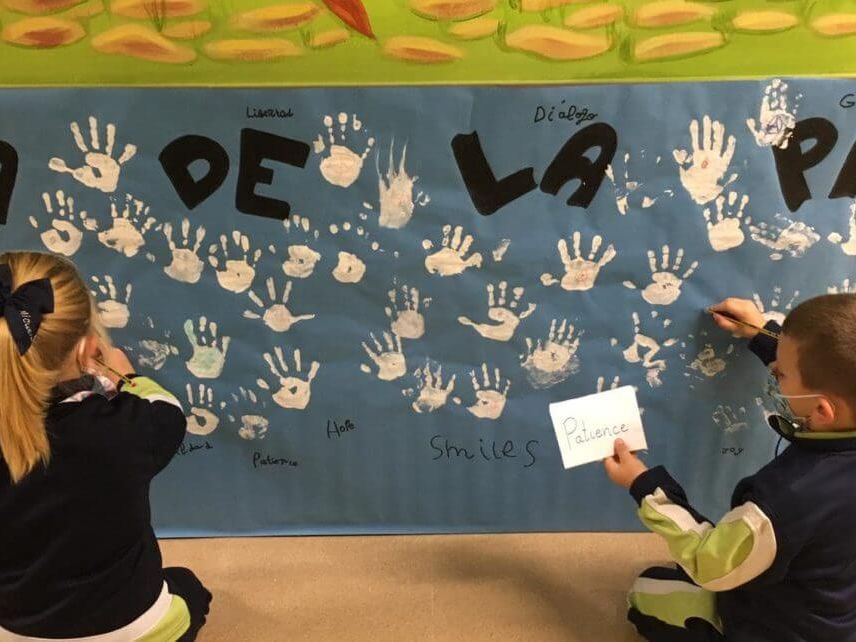 Dos alumnos escriben en un mural del Día de la Paz lleno de huellas de manos
