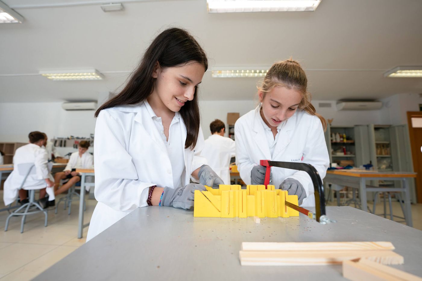 Dos alumnas de Secondary con bata blanca sierran una pieza en el aula taller