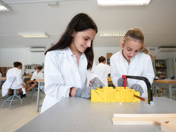 Dos alumnas de Secondary con bata blanca sierran una pieza en el aula taller