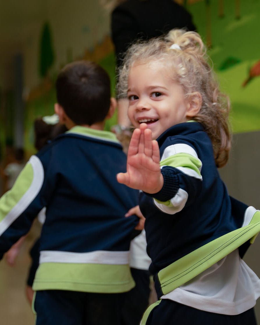 Una niña de Infants saluda mientras camina en fila por el pasillo