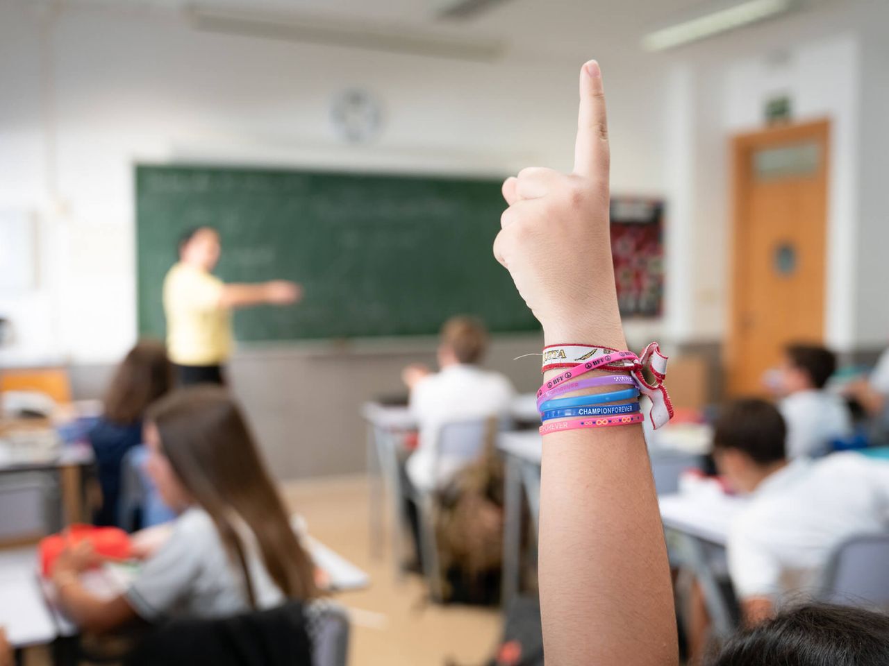 Una mano levantada en clase, con la profesora al fondo junto a la pizarra