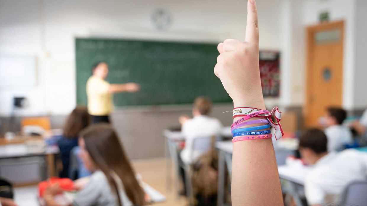 Una mano levantada en clase, con la profesora al fondo junto a la pizarra